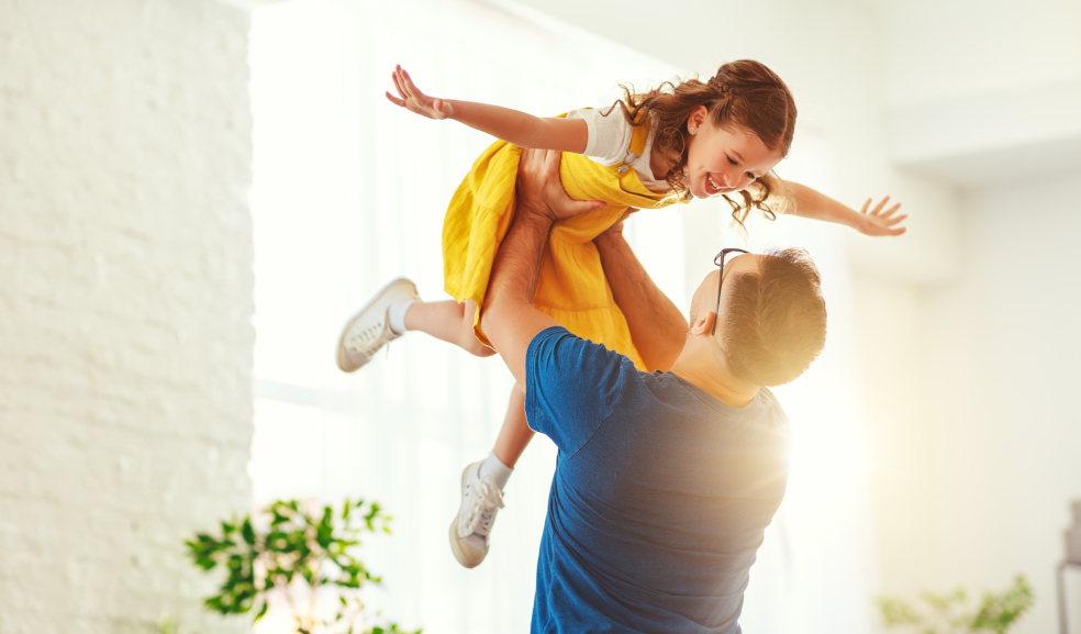 Das Bild zeigt einen Mann, der ein Kind in einem gelben Kleid spielerisch hochhebt. Das Kind streckt die Arme aus und wird in einer hell erleuchteten, freundlichen Umgebung gehalten. Beide Personen wirken fröhlich und es herrscht eine warme, familiäre Atmosphäre.