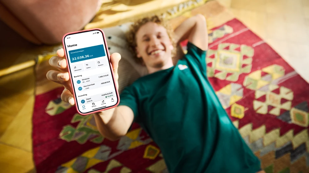 Ein junger Mann mit blonden Locken liegt auf einem rot gemusterten Teppich auf dem Boden und hält den Bildschirm seines Smartphones in die Kamera hoch. Auf dem Bildschirm sieht man die Consorsbank App.