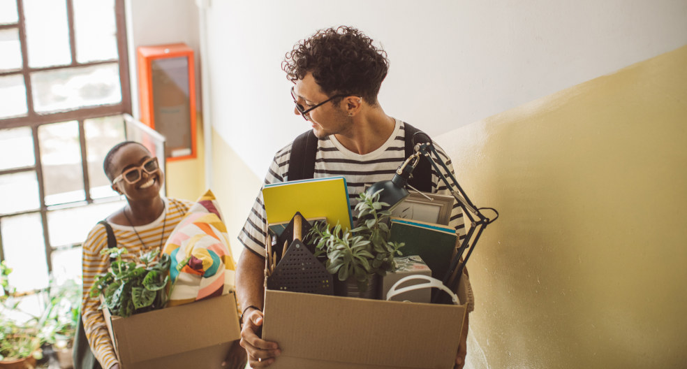Eine junge Frau und ein junger Mann mit Brille und kurzen braunen Haaren mit Umzugskisten im Treppenhaus