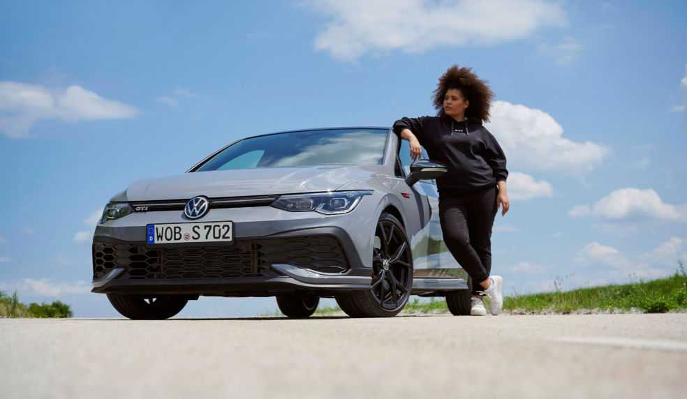 Frau mit dunklen Locken steht neben einem grauen Volkswagen GTI