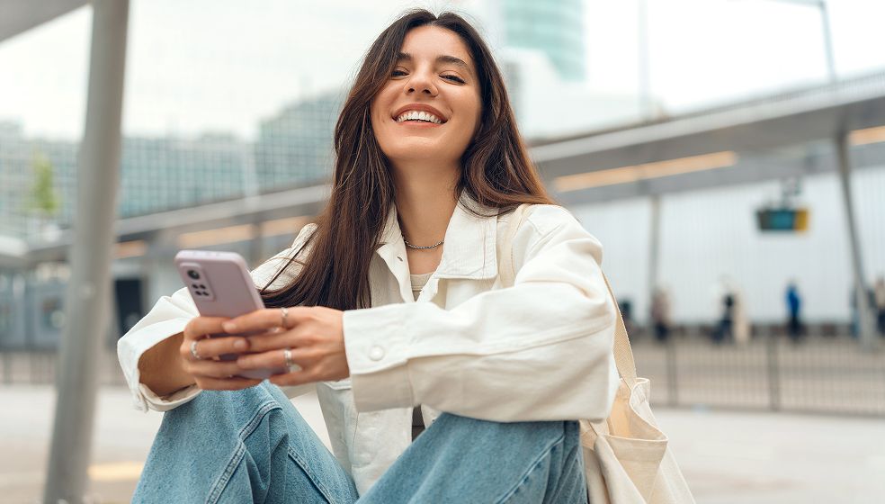 Eine junge Frau mit langen braunen Haaren sitzt draußen an einem modernen Bahnsteig, trägt eine helle Jacke und Jeans und hält ein Smartphone in der Hand. Im Hintergrund sind unscharfe Personen sowie Gebäude und Verkehrsanzeigen zu sehen.