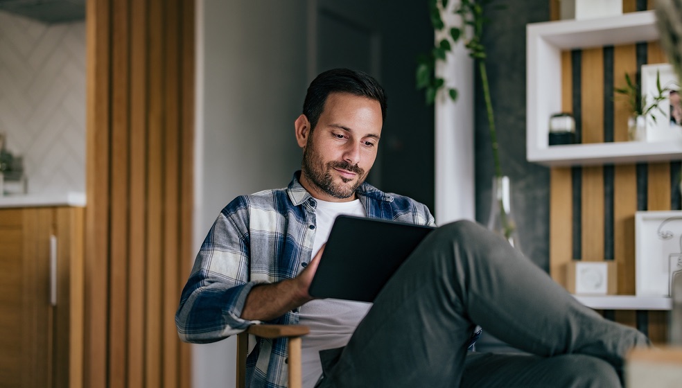 Dunkelhaariger Mann in Karohemd sitzt auf einem Stuhl und schaut in sein Tablet
