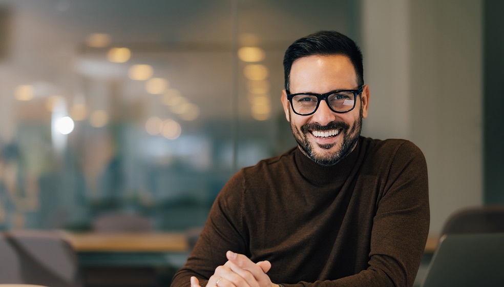 Mann mit brauen Haaren, Vollbart, Brille im braunen Pullover lächelt in die Kamera