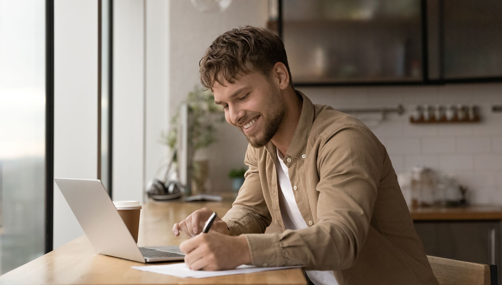 Ein Mann sitzt an einem Tisch in einer modernen Küche oder einem offenen Wohnraum. Vor ihm steht ein Laptop, daneben liegt ein Stift und ein Blatt Papier. Außerdem steht ein Kaffeebecher auf dem Tisch. Im Hintergrund sind Küchenschränke und einige Küchenutensilien zu sehen. Durch die großen Fenster fällt Tageslicht in den Raum.