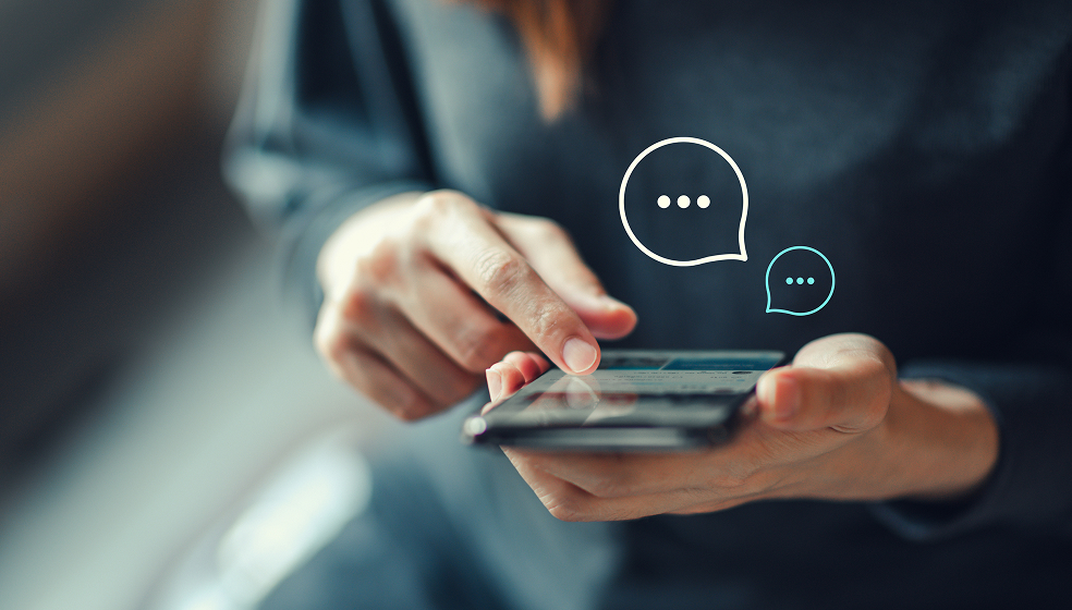 Das Bild zeigt eine Person, die ein Smartphone in der Hand hält und mit dem Finger auf den Bildschirm tippt. Über dem Smartphone sind zwei Sprechblasen mit drei Punkten als Symbol für eine Chat-Nachricht eingeblendet.