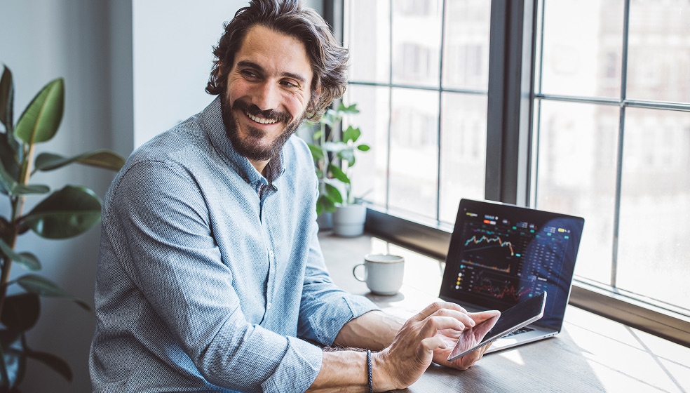 Das Bild zeigt eine Person, die an einem Schreibtisch vor einem großen Fenster sitzt. Auf dem Schreibtisch steht ein Laptop, dessen Bildschirm mehrere bunte Kurvendiagramme und Zahlen zeigt, vermutlich Finanzdaten oder Aktienkurse. Die Person hält ein Tablet und scheint darauf zu tippen. Im Hintergrund sind Pflanzen und eine Kaffeetasse zu sehen, die Szenerie wirkt wie ein modernes Büro oder Homeoffice.
