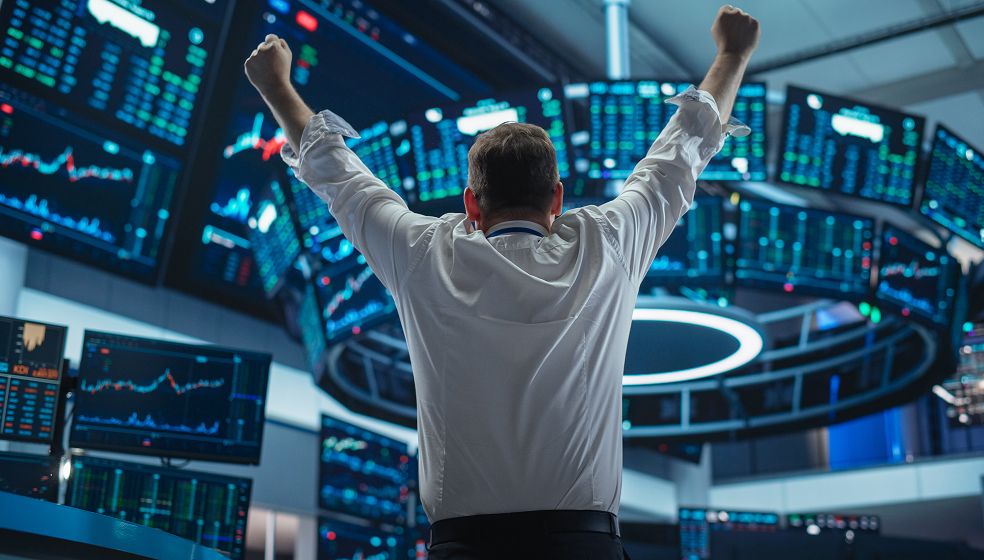 Bildbeschreibung: Ein Mann in Hemd steht mit erhobenen Armen vor zahlreichen großen Bildschirmen in einem modernen Handelsraum oder einer Börse. Die Bildschirme zeigen verschiedene Finanzdiagramme, Kursentwicklungen und Zahlen. Der Mann wirkt siegreich oder feiert einen Erfolg. Detaillierte Inhalte der Diagramme sind nicht lesbar.