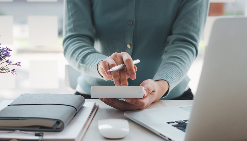 Blick auf den Schreibtisch mit Laptop und Journal hinter dem eine Frau steht, von der man nur die Hände sieht, die einen Taschenrechner in der Hand hält und etwas berechnet. 