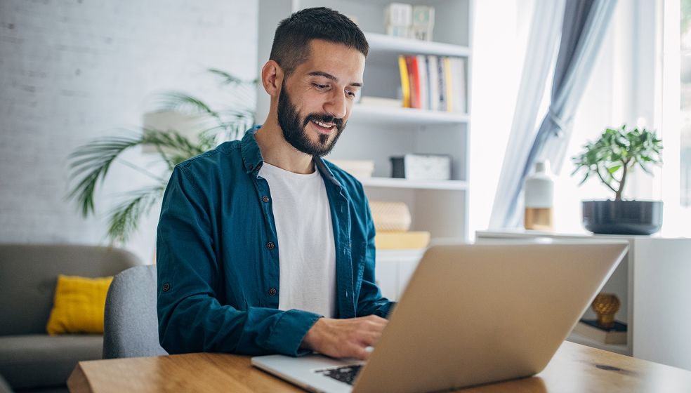 Das Bild zeigt eine Person, die an einem Schreibtisch sitzt und auf einem Laptop arbeitet. Im Hintergrund sind Regale mit Büchern, eine Pflanze und ein Fenster mit Vorhang zu sehen. Die Umgebung wirkt wie ein modernes, helles Homeoffice.