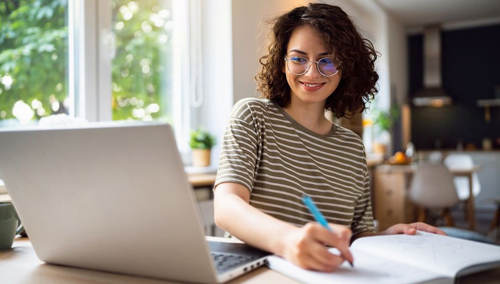 Eine Person sitzt an einem Tisch mit einem geöffneten Laptop und schreibt in ein Notizbuch. Im Hintergrund ist eine helle Wohnung mit Fenstern und Kücheneinrichtung zu sehen.