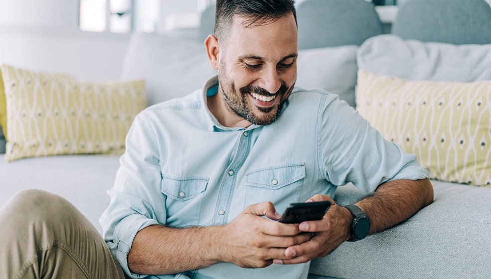 Mann mit braunen Haaren im blauen Jeansheamd lehnt lässig vor seiner Couch, hält ein Smartphone in der Hand und blickt lächelnd auf den Bildschirm