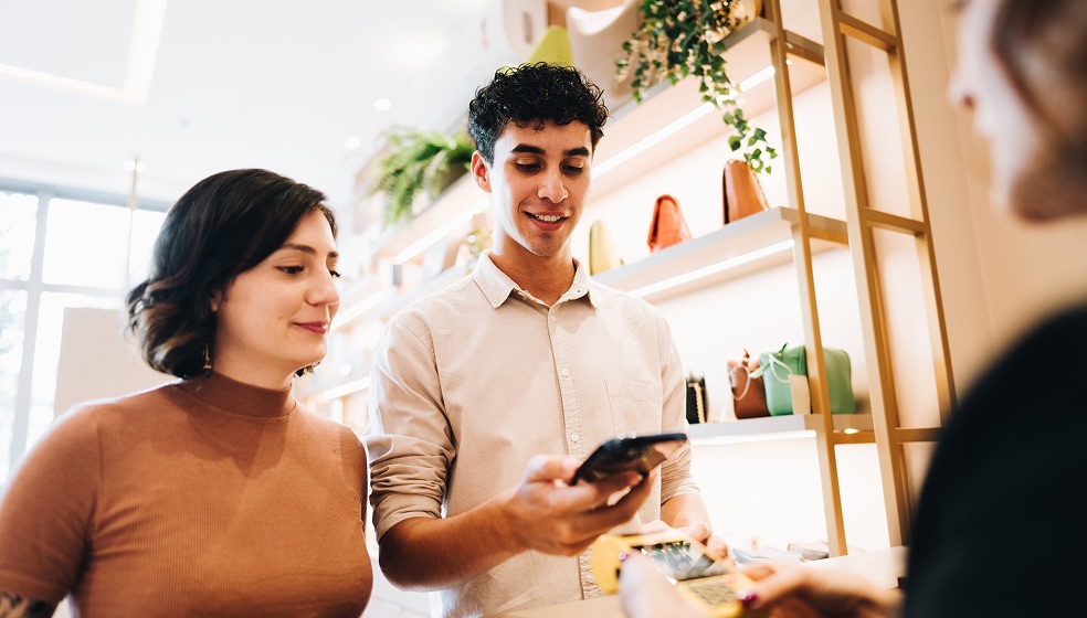 Ein Mann und eine Frau mit dunklen Haaren stehen vor einer Kassiererin und ihrem Terminal. Der Mann hat sein Smartphone in der Hand. 