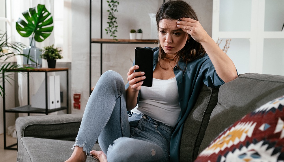 Frau mit dunklen Haaren und Pferdeschwanz sitzt auf einer Couch, hält ein Smartphone in der Hand und blickt verzweifelt auf den Bildschirm
