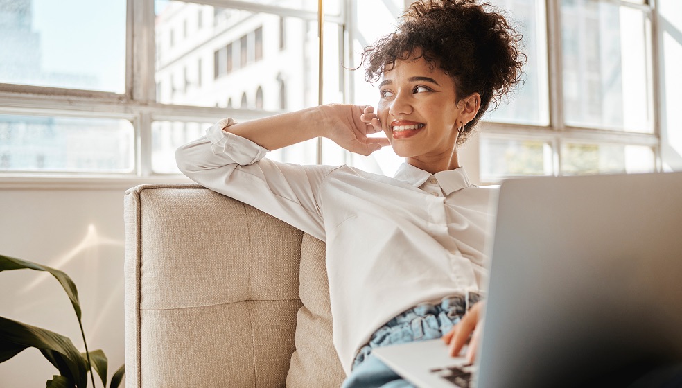 Frau mit Afro-Frisur sitzt entspannt auf einem Sofa, hat ein Notebook auf dem Schoß und blickt lächelnd zur Seite