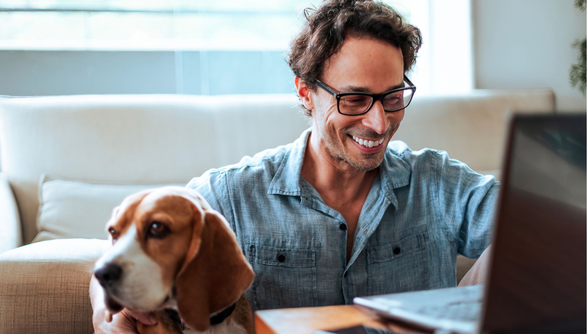 Eine Person sitzt auf einem Sofa zu Hause an einem Laptop. Neben der Person sitzt ein Hund, vermutlich ein Beagle. Die Szene wirkt entspannt und häuslich.