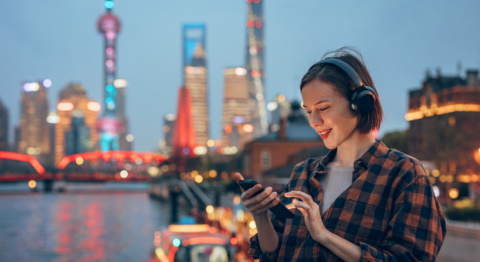 Die Aufnahme zeigt eine Person mit Kopfhörern und Mobiltelefon am Ufer eines Flusses in einer modernen Großstadt bei Dämmerung. Im Hintergrund sind beleuchtete Hochhäuser, Brücken und reflektierende Stadtlichter auf dem Wasser zu sehen.