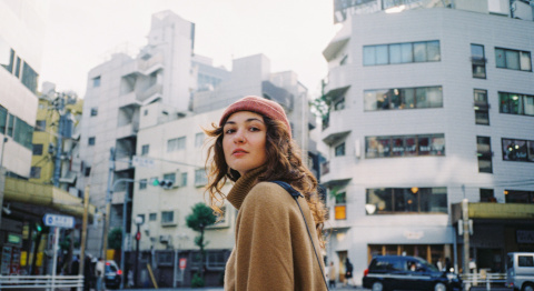 Eine Person mit langen Haaren und einer roten Mütze steht auf einer Straße in einer städtischen Umgebung, umgeben von mehreren mehrstöckigen Gebäuden und vorbeifahrenden Autos. Die Atmosphäre wirkt belebt und urban.