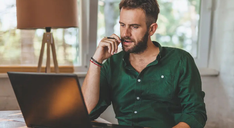 Mann mit kurzen blonden Haaren und grünem Hemd sitzt an einem Schreibtisch und blickt nachdenklich auf seinen Laptop. 