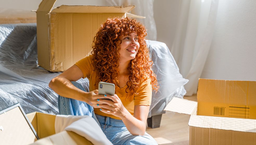 Eine Person mit roten, lockigen Haaren sitzt in einem hellen Zimmer auf dem Boden zwischen mehreren geöffneten Umzugskartons. Die Person trägt ein gelbes T-Shirt und Jeans und hält ein Smartphone in der Hand. Im Hintergrund ist ein mit Folie abgedecktes Möbelstück zu sehen. Licht fällt durch ein Fenster von rechts ins Zimmer.