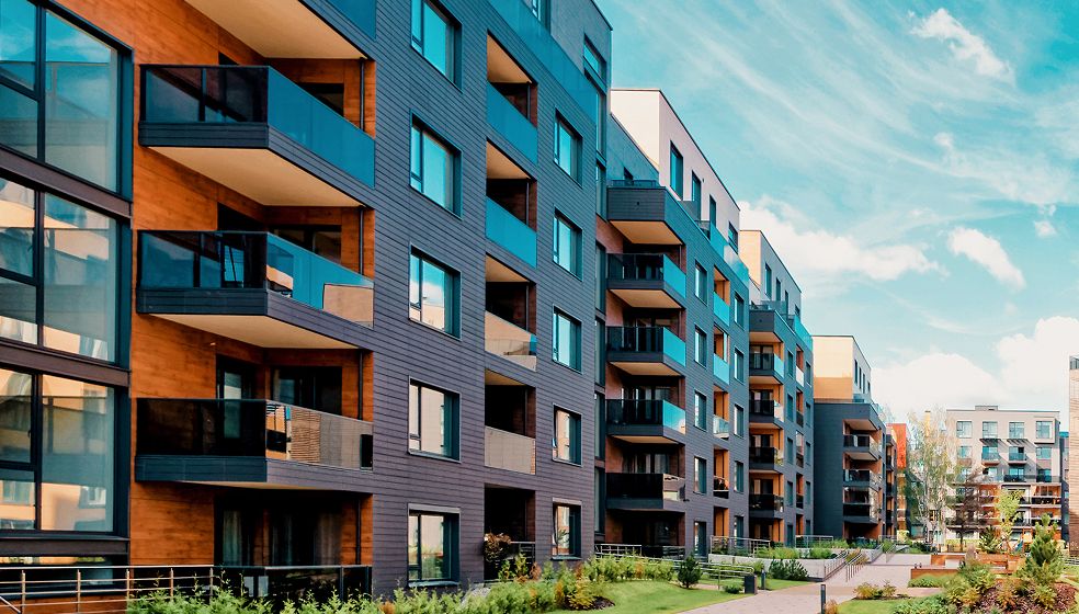 Das Bild zeigt moderne, mehrstöckige Wohngebäude mit großen Fenstern und Balkonen. Im Vordergrund befinden sich grüne Pflanzen und ein Gehweg, der zwischen den Gebäuden verläuft. Die Fassade der Häuser ist abwechselnd in Holz- und Grautönen gestaltet. Der Himmel ist blau mit einigen Wolken.