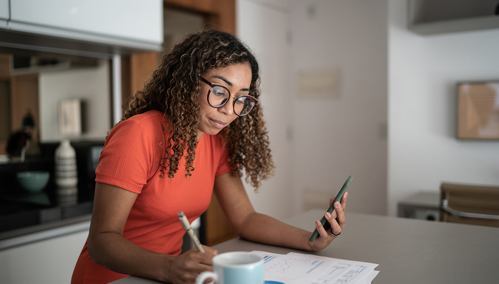 Eine Person mit lockigen Haaren trägt ein orangefarbenes Kleid und befindet sich in einer modernen Küche. Sie steht an einer Kücheninsel, hält in der einen Hand ein Smartphone und schreibt mit der anderen Hand auf einem Blatt Papier mit Diagrammen. Auf dem Tisch stehen außerdem eine Tasse und einige Unterlagen mit grafischen Darstellungen.