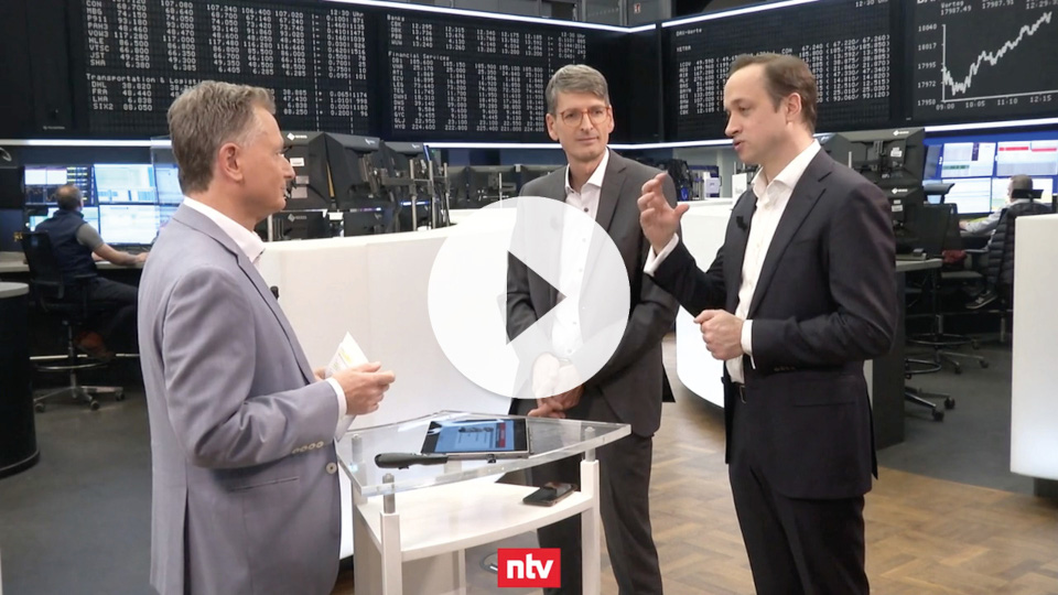 Drei Männer in Anzügen stehen in einer Börse vor verschiedenen Monitoren und diskutieren miteinander