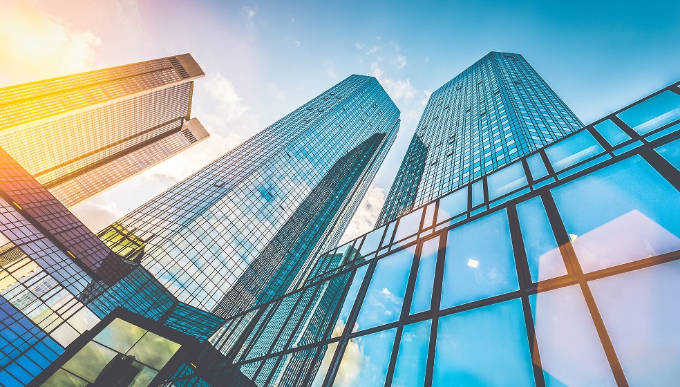 Blick auf mehrere komplett verglaste hohe Bürogebäude, die in der Abendsonne in gelb, rot und blautönen schimmern. 