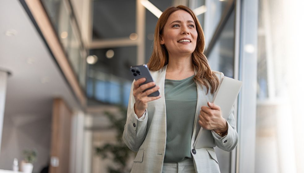 Frau mit langen braunen Haaren in grauem Blazer steht vor einem Bürogebäude, hält ein Smartphone in der Hand und blickt lachend zur Seite