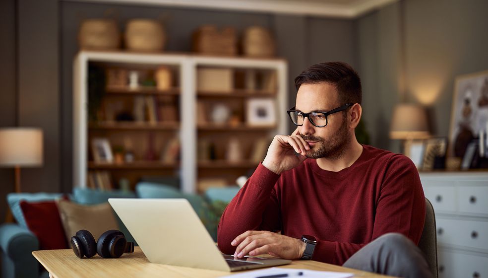 Eine Person sitzt an einem Schreibtisch in einem Wohnzimmer und arbeitet an einem Laptop. Auf dem Tisch liegen Kopfhörer und einige Papierunterlagen. Im Hintergrund sind ein Sofa, Regale mit Büchern und Dekoration sowie zwei Lampen zu sehen. Die Atmosphäre wirkt ruhig und häuslich.