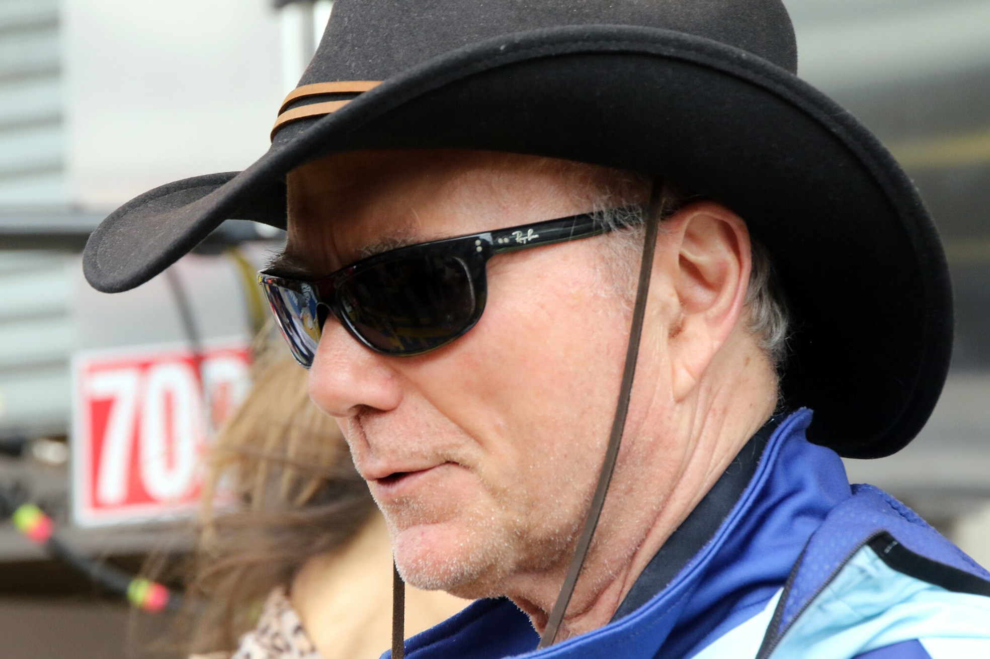 Das Foto zeigt eine Person mit grauen Haaren, die einen schwarzen Cowboyhut und eine blau-schwarze Jacke trägt. Im Hintergrund ist ein Schild mit der Zahl 703 zu sehen.