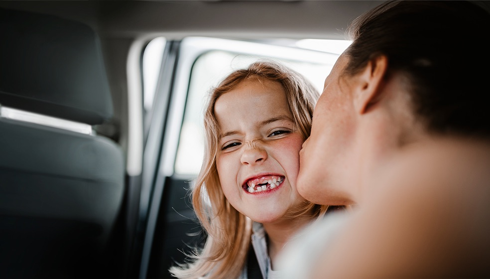 Eine Mama küsst ihre Tochter auf der Rückbank eines Autos auf die Wange. Das Mädchen hat lockige lange braune Haare, grinst und man sieht eine Zahnlücke