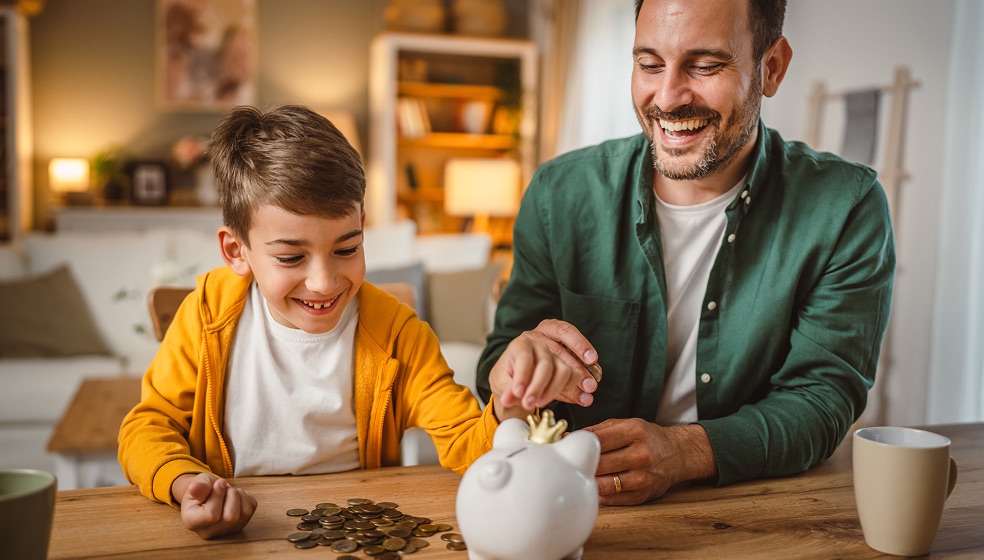Papa und Sohn befüllen gemeinsam ein weißes Porzellansparschwein mit Kleingeld
