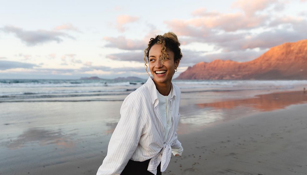 Eine Person steht am Strand bei Sonnenuntergang. Im Hintergrund sind das Meer mit kleinen Wellen und Berge zu sehen, die in warmes Abendlicht getaucht sind. Die Person trägt ein helles, langärmliges Oberteil.