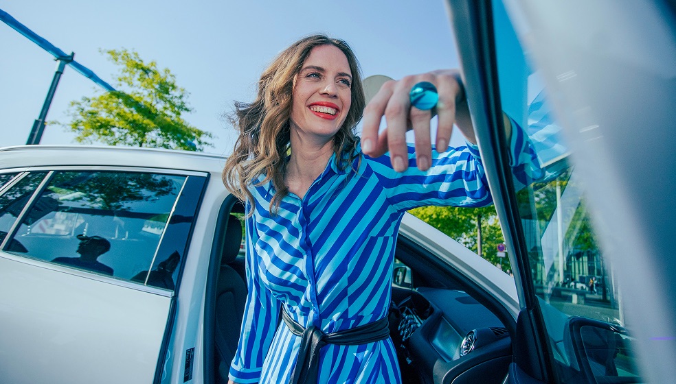 Frau mit langen lockigen rotblonden Haaren im blau-gestreiften Kleid lehnt lachend in einer Autotür 