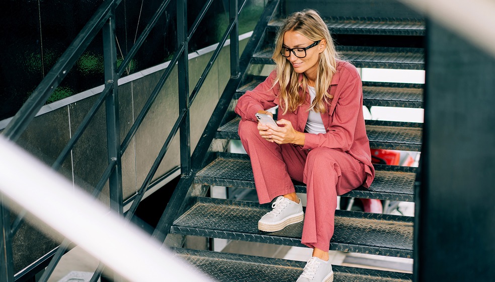 Blonde Frau in legere pinkem Anzug sitzt auf einer Treppe und blickt lächelnd auf ihr Smartphone
