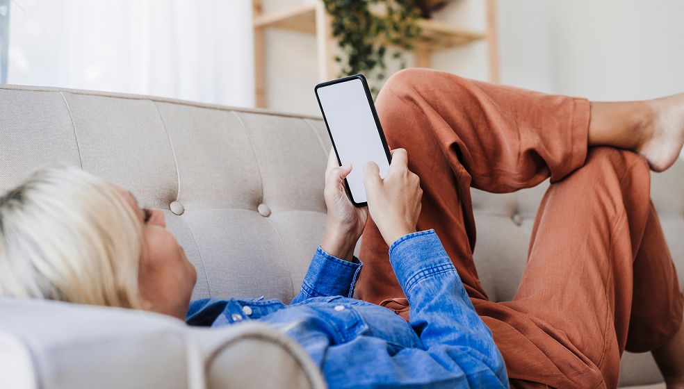 Das Bild zeigt eine Person, die entspannt auf einem Sofa liegt und ein Smartphone in der Hand hält. Die Person trägt ein blaues Hemd und rotbraune Hosen. Der Bildschirm des Smartphones ist leer. Das Gesicht der Person ist nur teilweise erkennbar. Im Hintergrund ist unscharf ein Regal mit einer Pflanze zu sehen.