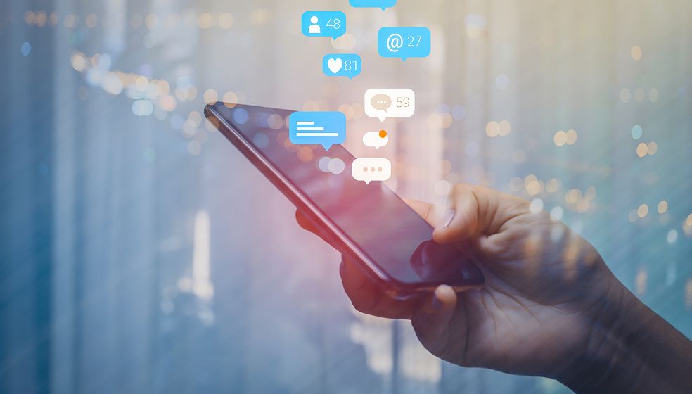 Das Bild zeigt eine Hand, die ein Smartphone hält. Über dem Smartphone erscheinen verschiedene Symbole für soziale Interaktionen, wie ein Benachrichtigungssymbol mit der Zahl 27, ein Herz mit der Zahl 81, ein Symbol für Nachrichten mit der Zahl 59 und ein Benutzersymbol mit der Zahl 48. Die Symbole sind in hellen Farben gehalten und symbolisieren neue Nachrichten, Likes, Kommentare und Follower. Der Hintergrund ist unscharf und zeigt Lichtpunkte, die eine moderne, digitale Umgebung andeuten.