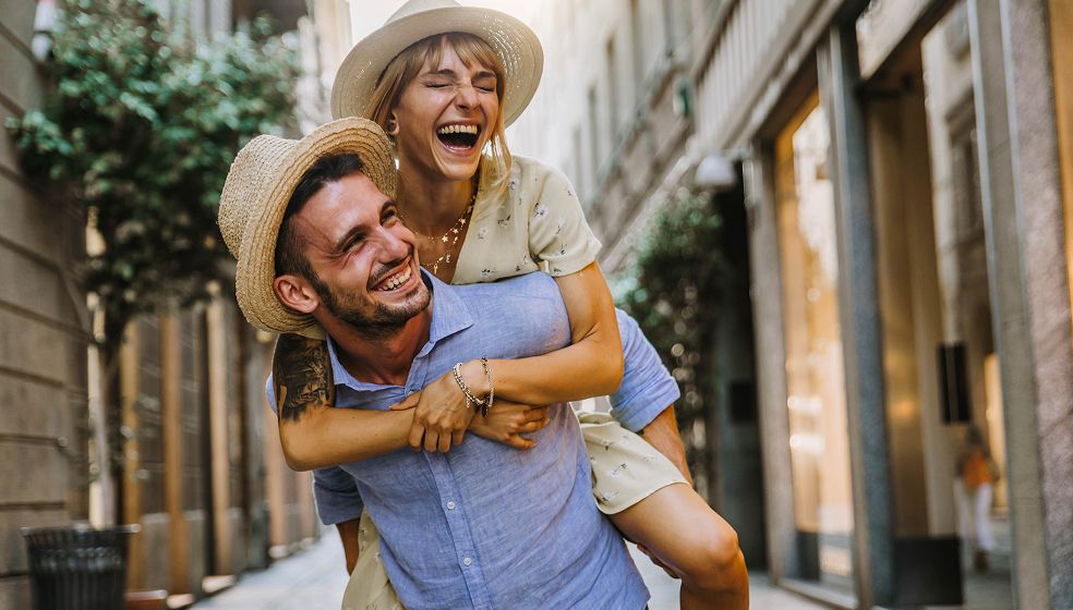 Das Bild zeigt zwei erwachsene Personen, die in einer sonnigen Stadtstraße unterwegs sind. Eine Person trägt einen Strohhut und ein hellblaues Hemd und trägt die andere Person auf dem Rücken. Die getragene Person trägt ein helles Sommerkleid und ebenfalls einen Hut. Beide wirken fröhlich und lächeln. Im Hintergrund sind Bäume, Gebäude und Schaufenster zu sehen.