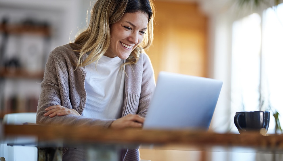 Frau mit langen blonden Haaren sitzt an einem Tisch mit ihrem Notebook und blickt lächelnd auf den Bildschirm