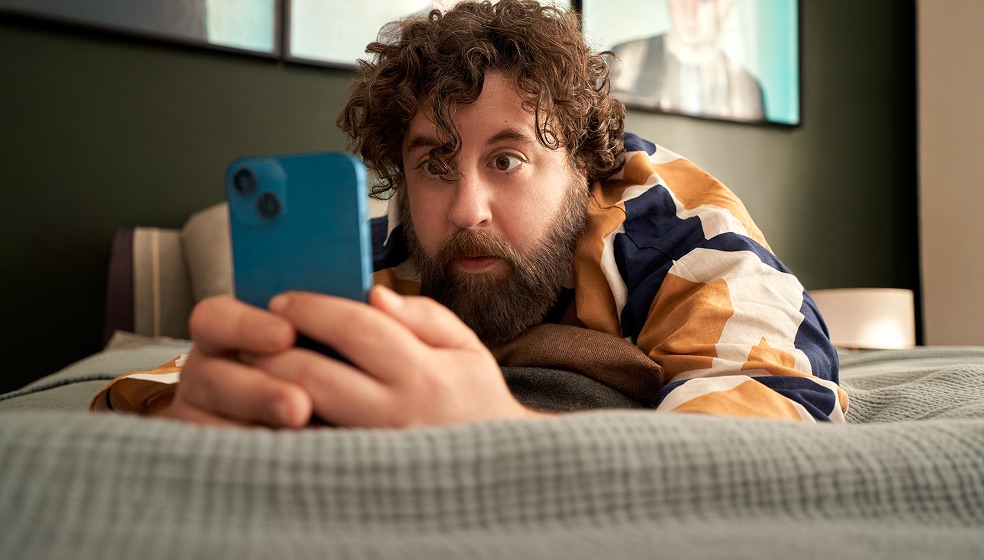 Mann mit braunen Locken und Vollbart liegt bäuchlings auf einem Bett, hält seine Hände mit einem Smartphone vor sich und blickt erstaunt auf den Bildschirm