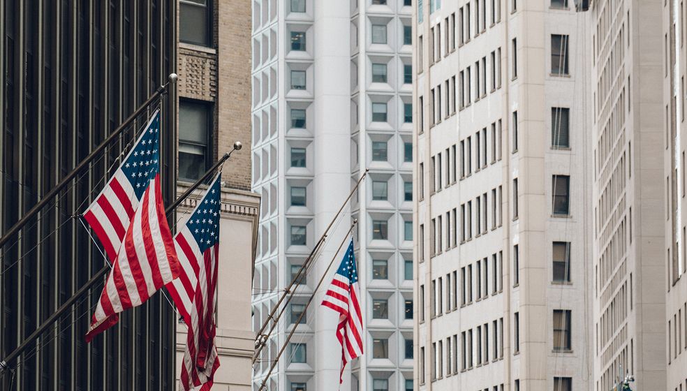 Das Bild zeigt mehrere amerikanische Flaggen, die an Stangen vor Hochhäusern in einer Stadt befestigt sind. Die Gebäude haben viele Fenster und eine moderne Fassade. Es sind keine Menschen zu sehen.