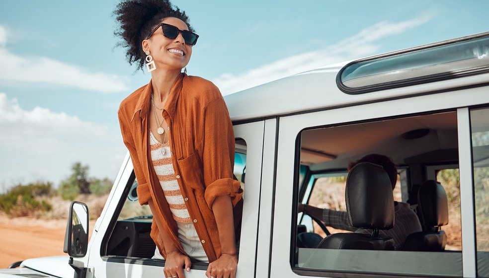 Frau mit lockigen dunkelbraunen Haaren, einer rostbraunen Bluse und Sonnenbrille lehnt sich lässig auf der Fahrerkabine eines Jeeps, der auf einer roten Sandstraße fährt. Am Straßenrand ist Busch zu erkennen.  