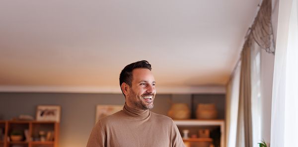 Das Bild zeigt einen Mann mit dunklen Haaren in einem hellbraunen Rollkragenpullover, der in einem Wohnzimmer mit Regalen, Bildern und Körben im Hintergrund steht. Rechts im Bild sind bodentiefe Fenster mit Vorhängen zu sehen, durch die Tageslicht einfällt.