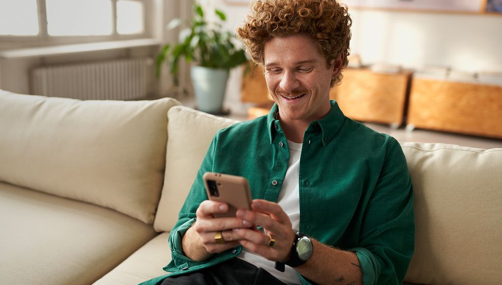 Eine Person mit lockigen Haaren sitzt auf einem beigen Sofa in einem hellen Raum und hält ein Smartphone in beiden Händen. Im Hintergrund sind ein grüner Blumentopf und Holzmöbel zu sehen.