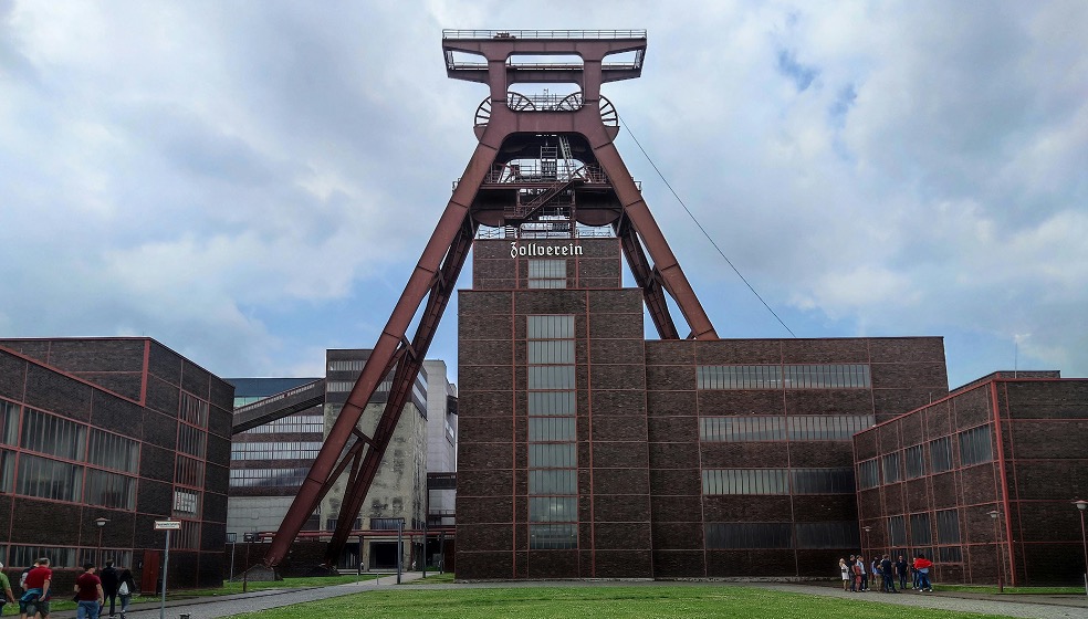  Blick auf die Gebäude der Zeche Zollverein im Ruhrgebiet mit Backssteingebäuden und einem rostroten Förderturm in der Mitte