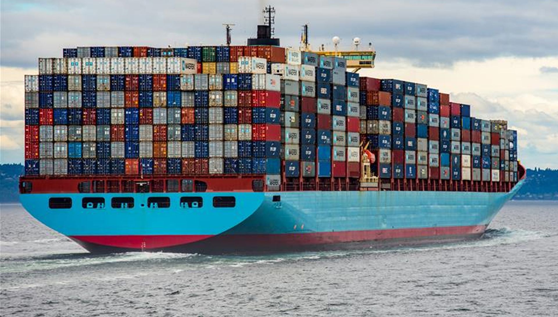 Das Bild zeigt ein großes Containerschiff, das auf dem Meer fährt. Das Schiff ist mit vielen, unterschiedlich farbigen Containern beladen, die gestapelt sind. Das Schiff ist vorne hellblau und rot. Im Hintergrund ist Wasser und bewölkter Himmel zu sehen.