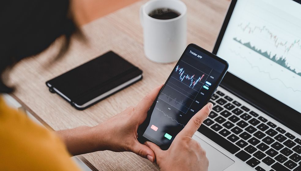 Eine Person sitzt an einem Schreibtisch und hält ein Smartphone in der Hand, auf dem eine Handels-App mit einem Diagramm angezeigt wird. Das Diagramm zeigt eine Kurve mit Schwankungen und steigender Tendenz. Im Hintergrund sind ein aufgeklappter Laptop mit einem ähnlichen Liniendiagramm, ein Notizbuch und eine Tasse Kaffee zu sehen. Zahlen- und Achsenbeschriftungen sind auf den Diagrammen nicht deutlich lesbar.