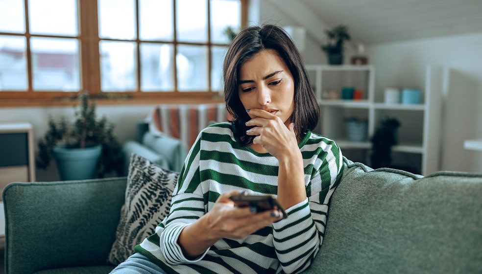 Frau mit halblangen dunkelbraunen Haaren sitzt auf einer grünen Couch und blickt nachdenklich auf ihr Smartphone, dass sie in der Hand hält