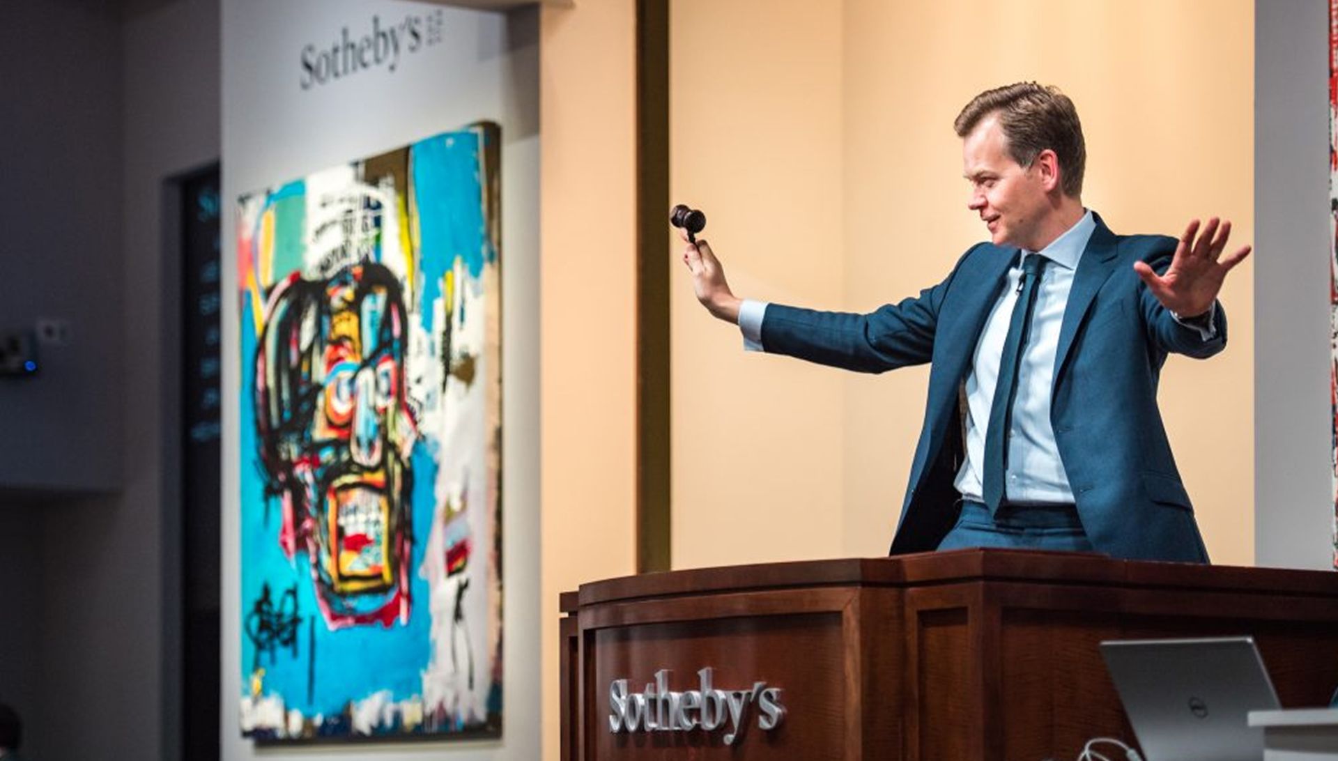 Das Bild zeigt eine Kunstauktion bei Sotheby’s. Im Vordergrund steht ein Auktionator im blauen Anzug hinter einem Rednerpult mit einem Auktionshammer in der Hand. Links im Hintergrund ist ein farbenfrohes Gemälde im Stil von Jean-Michel Basquiat zu sehen. Auf dem Pult steht deutlich sichtbar „Sotheby’s“.