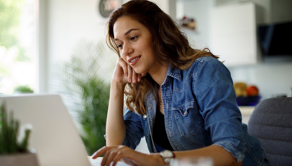 Frau mit braunen lockigen Haaren sitzt an einem Schreibtisch, hat ein Notebook vor sich und blickt nachdenklich lächelnd auf den Bildschirm 